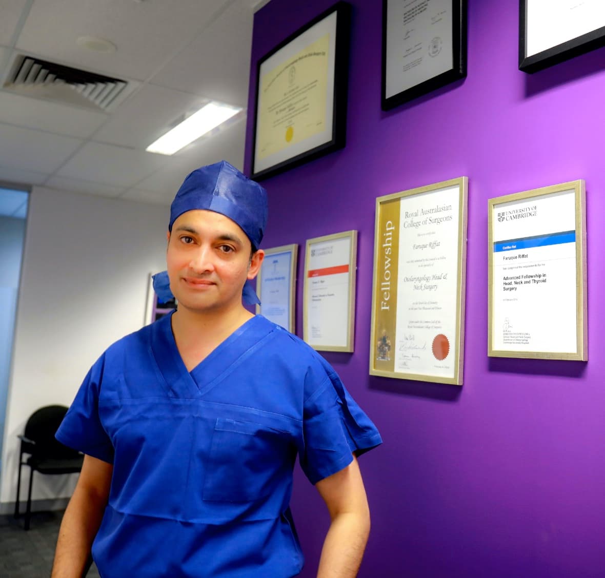 A/Prof Faruque Riffat in scrubs in front of his framed fellowship certificates.