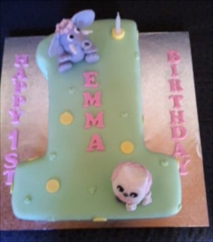 Number-1 shaped 1st birthday cake in sage and pink with an elephant figurine, reading 'Happy 1st Birthday Emma'